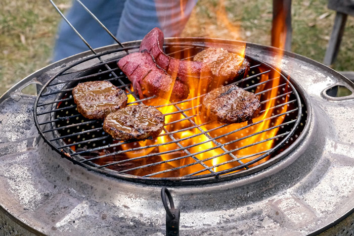 Burgers and steak cooking on an outdoor fire pit.
