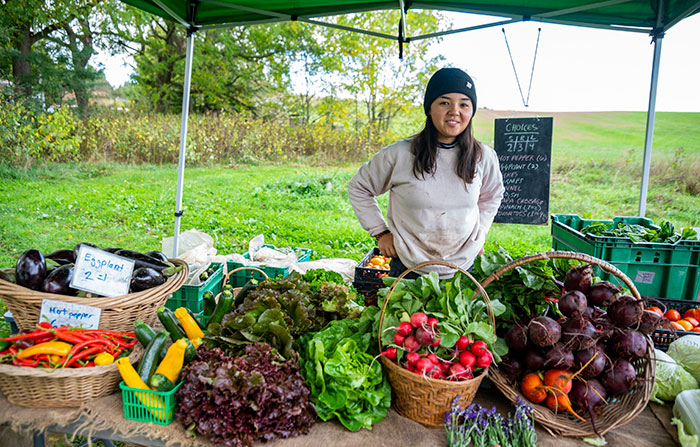 A vendor stands under a tent, which shelters their stand at a farmer's market.