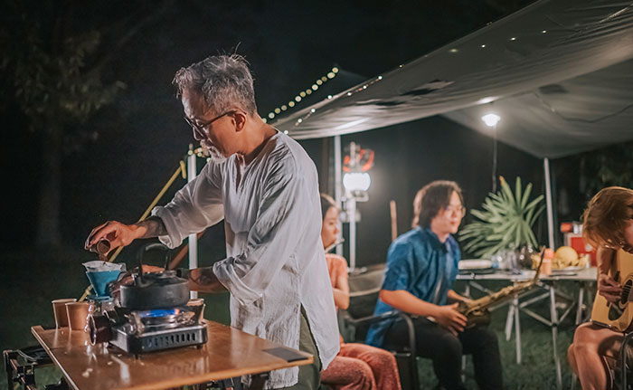 A man prepares drinks, behind him there is a social gathering under a portable canopy.