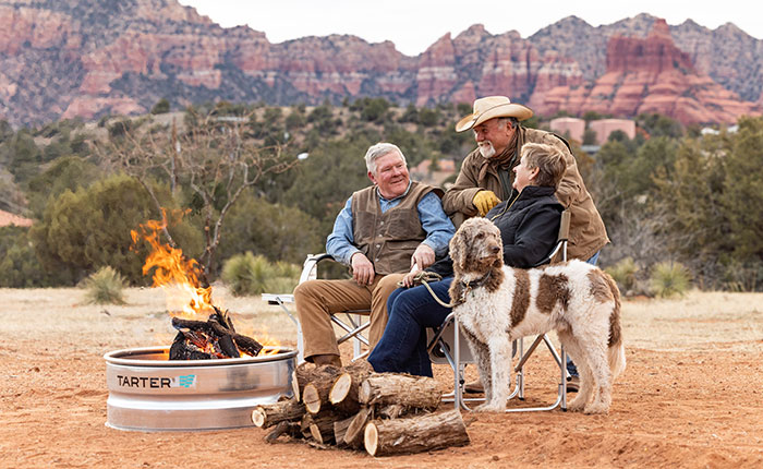 A group of friends and a dog sit by a fire ring, in a desert setting.